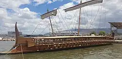 Color photograph of a long wooden sailing ship at anchor, with two folded masts, an upper deck, a middle deck and three oarsmen's decks.