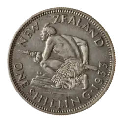 A coin featuring a crouched Māori atop a rocky escarpment. He holds a taiaha war-staff with both hands pointing it and facing forward off the escarpment. He is muscular and shirtless, but wears a piupiu skirt.