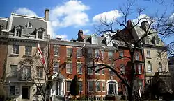 Depiction of street with red townhouses connected together, each with white window shutters. Blue sky visible above with some cloud wisps. Tree without leaves in the right foreground of the picture. White-and-red flag, presumably of a country, hung on the far left of the picture.