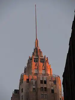 The spire as seen at sunset. The spire is made of metal and stands above a series of stone setbacks.