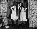 A black and white photograph of two women, both wearing white knee-length dresses, black tights, black shoes, and white Venetian masks covering their eyes and nose. The women have cigarettes in their mouths and are holding the tips to each other.