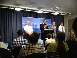 In a small conference room, a seated audience listens to Angus Houston speaking behind a podium and flanked by three other officials.