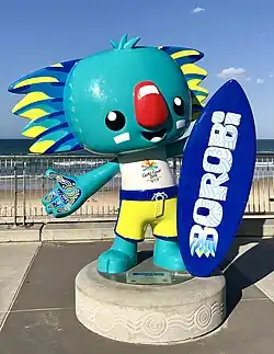A statue of a cyan koala with blue and yellow fur details and a red nose. He is holding a blue surfboard with the word "Borobi" written on it.