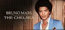 Bruno Mars wearing a blue jacket and a white shirt, posing in front of a wooden wall