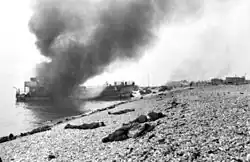 burning ship at sea with smoke billowing up, at least 20 to 30 dead lying on the beach