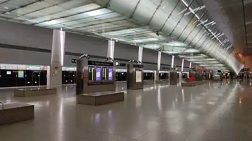 A large island platform which includes flight information displays. An illuminated linkbridge spans over the platform.