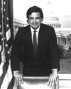 A Latino man leans over a chair and smiles at the camera, with the U.S. Capitol in the background