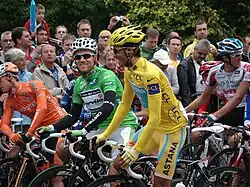 Several cyclists sit on their bicycles. In the center of the image are two, wearing solid yellow and green jerseys. Spectators watch in the background.