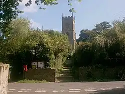 Square church tower surrounded by trees with a road in the foreground.