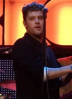 A twenty-six year-old man is shown in three-quarter view. He is standing centre-stage, half-turned with hands clapped together. He is partly obscured by a microphone stand in front. Behind him is a drummer at his kit. Beyond that is the lighting, which includes the partly spelled letters R-O-C-K-C-O-R in large orange-yellow.