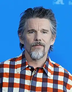 A Caucasian male with brown hair, wearing a two-piece dark gray suit with a white shirt.