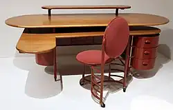 Image of two pieces of furniture that were made for the Johnson Administration Building. There is a desk with an oval wooden surface. In front of it is a three-legged red chair.