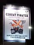 An advertising billboard of Galeria Katowicka shopping mall that means "Black Friday. Buy something and get a bag with a graphic designed by Roobens"