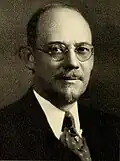 White male with dark hair and a thin beard in a dark suit