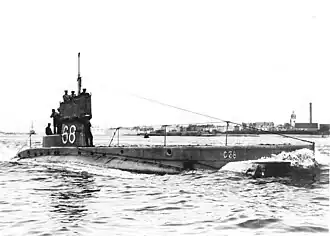 Black and white photograph of a C-class submarine surfacing in port. Five men are visible standing on the rear of the vessel