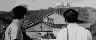 A picture showing two detectives looking up at Gondo's house on the hill, with an array of smaller rickety houses in the foreground
