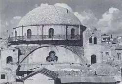 A large domed roof sits upon a bricked parapet, which includes arched windows, three of which are visible. The base of the parapet is surrounded by a narrow veranda, which is fenced with decorative metal grating. Below this, a central clover shaped window lies at the top of an arched wall. Two supporting towers stand on the left and right, each containing an arched window. The right tower, capped with a small dome, is a storey higher, reaching to the top of the bricked parapet, which supports the domed roof.
