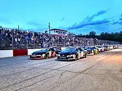 A photograph of the frontstretch of Greenville-Pickens Speedway. Cars are parked and lined up in a double-wide formation.