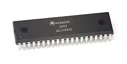 Close up of a small black rectangular microprocessor chip with small metallic prongs extending from the side downward.