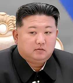 An official image of Kim Jong Un. He is wearing a black striped suit and standing in front of a flag.