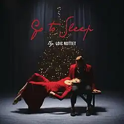 Shot of Nottet and a female laying over a bench in front of a Christmas tree; information on the song is superimposed on them.