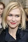 Mugshot of a woman with blonde hair and grey eyes. She is smiling; we can see her dimples.