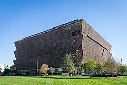 A three tiered golden colored building with no visible windows and a green lawn in front.
