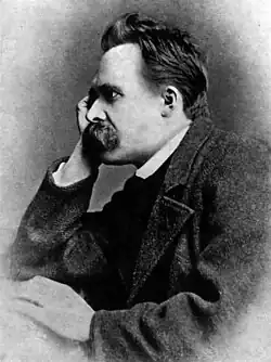 Black-and-white photo showing a man with a thick mustache from the side, wearing a dark suit and resting is his head on his hand