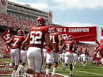 Football players run onto a football field in two rows under a crimson banner.