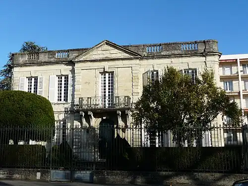 One-storey high building with 5 high windows, with a railing on its flat roof, preceded by a terrace and a fence.