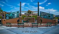 A brick plaza with three benches and a wall of plaques. Sitting in one of the benches is a bronze statue of Calhoun.