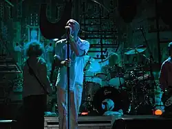 Male singer in white shirtsleeves and trousers, with a band behind him on a small stage.