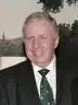 Head and shoulders of Paul Murphy, stood in a dark suit with a shirt and a green tie, in front of a white wall.