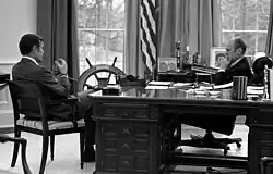 Gerald Ford sits behind the Wilson Desk talking with George H. W. Bush who sits beside it
