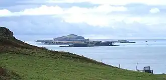 Brown-coloured islands of varying elevations lie in a calm sea beneath cloudy skies beyond a grassy slope.