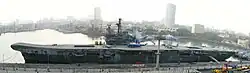 A small aircraft carrier tied up at a wharf. A helicopter and two aircraft sit on the flight deck. Other ships and skyscrapers are in the background.