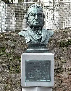 An outdoor bust of a bearded man wearing a large patterned cap and a dicky bow tie. A plaque beneath reads: Vincent Wallace composer. Born Waterford 1812, died Pyrenees 1865. "In happy moments day by day, The sands of life may pass. In swift but tranquil tide away, From times inferring glass."