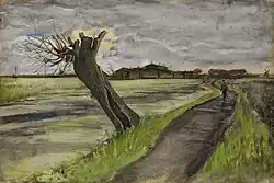 A dead pollard willow hangs over a pond. A path on the right leads to some large buildings on the horizon with a windmill on the right. A man is walking down the path.