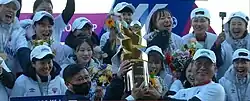 Smiling players from Incheon Hyundai Red Angels women's football team, along with their coach and chairman, lift a large golden grophy.
