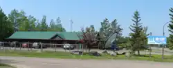 A photograph of the WTIP broadcasting station taken on a sunny day. The brown building is long and low (likely one story) with a green corrugated metal roof. There is a large satellite dish in front, as well as an antenna behind the building. A sign reads, "WTIP Community Radio wtip.org 90.7 FM." There is also a parking lot around the building with several vehicles.