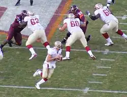 A quarterback rolls out to throw a pass while three linemen block their opponents at the line of scrimmage.