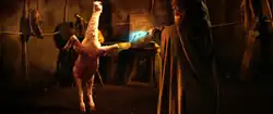 A little man in a hooded cloak with his back to the camera holds a lightening wand toward a two-legged animal that appears to be part goat and part ostrich.