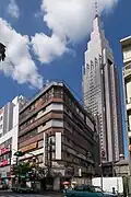 In the foreground: the abandoned Yoyogi Kaikan [ja] building that appears in the film. The demolition of the building was completed in January 2020 and it no longer exists. Note that the rooftop shrine in this film is fictional. Back: the NTT DoCoMo Yoyogi Building (Docomo Tower), which frequently appears in Shinkai's films.